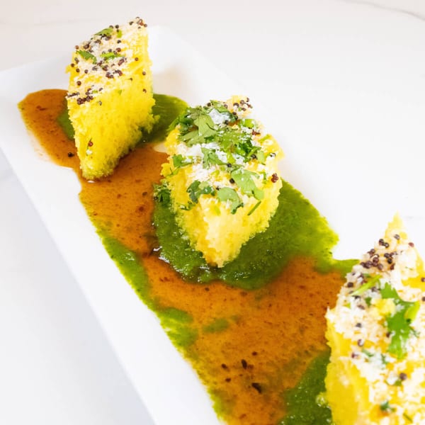 Three pieces of yellow Indian dhokla, garnished with cilantro and spices, on a white plate with green and brown sauces.