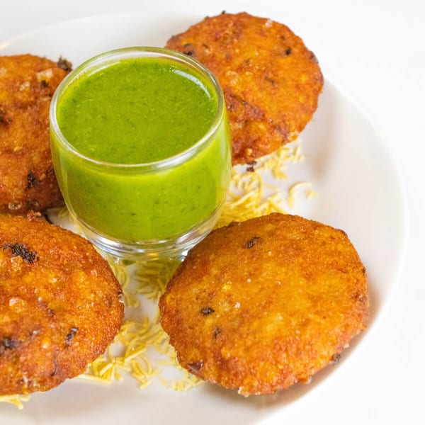 Four golden-brown Indian cutlets are arranged around a glass of vibrant green chutney on a white plate.