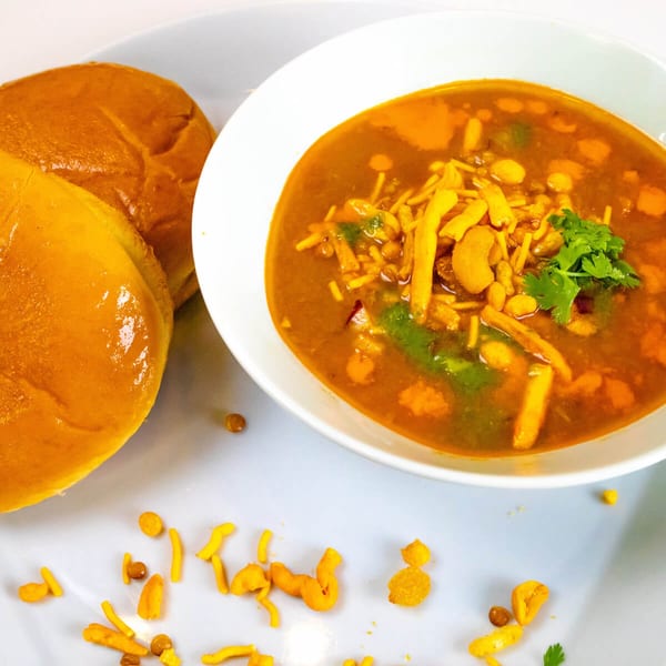 A white bowl of Indian misal pav, a spicy curry, with two pav buns on a white plate.